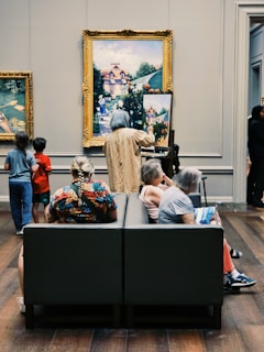 A gallery setting where an artist copies a large framed painting exhibited on the wall. The artist is standing in front of the painting, using an easel to recreate the artwork on a smaller canvas. In the foreground, several people are seated on a bench, observing the art and artist. The room has wooden floors and the environment exudes a contemplative atmosphere.