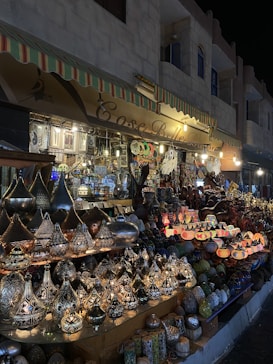 A market stall displaying a variety of ornate metal lamps and decorative items. The lamps feature intricate patterns and are arranged in rows, illuminated to highlight their detailed craftsmanship. The surrounding area showcases a colorful array of additional crafts, creating a vibrant and lively atmosphere.
