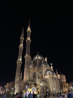 An ornate mosque with large domes and two tall minarets rises majestically against the night sky, illuminated by strategic lighting. The intricate architectural details display Islamic art influences. People gather around the building, enjoying the evening atmosphere.