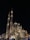 An ornate mosque with large domes and two tall minarets rises majestically against the night sky, illuminated by strategic lighting. The intricate architectural details display Islamic art influences. People gather around the building, enjoying the evening atmosphere.