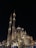 An ornate mosque with large domes and two tall minarets rises majestically against the night sky, illuminated by strategic lighting. The intricate architectural details display Islamic art influences. People gather around the building, enjoying the evening atmosphere.