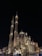 An ornate mosque with large domes and two tall minarets rises majestically against the night sky, illuminated by strategic lighting. The intricate architectural details display Islamic art influences. People gather around the building, enjoying the evening atmosphere.