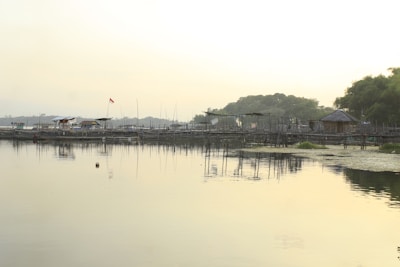 A serene lakeside scene with calm water reflecting the sky and surrounding structures. Wooden huts and structures line the shore, with a few trees and greenery. An Indonesian flag is flying on one of the structures.