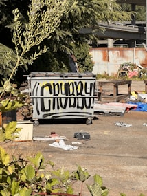 A large gray dumpster is positioned in an outdoor area surrounded by overgrown plants and debris. The front of the dumpster is spray-painted with the word 'CRUBBY!' in bold black letters. The background includes a rusted corrugated metal fence and an elevated roadway. Various discarded items and trash are scattered across the ground, adding to the scene's disordered appearance.