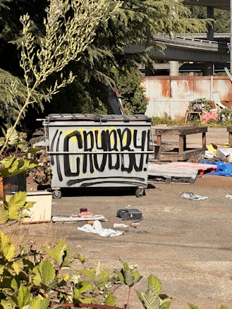 A large gray dumpster is positioned in an outdoor area surrounded by overgrown plants and debris. The front of the dumpster is spray-painted with the word 'CRUBBY!' in bold black letters. The background includes a rusted corrugated metal fence and an elevated roadway. Various discarded items and trash are scattered across the ground, adding to the scene's disordered appearance.