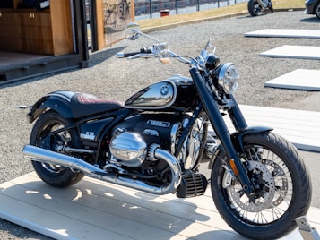 A sleek motorcycle is displayed on a light wooden platform outdoors. The motorcycle has a glossy black finish with chrome details, including the exhaust pipes and engine covers. The logo on the fuel tank is distinguishable. It is parked in an area with gravel ground and wooden platforms around, with a modern structure visible in the background.