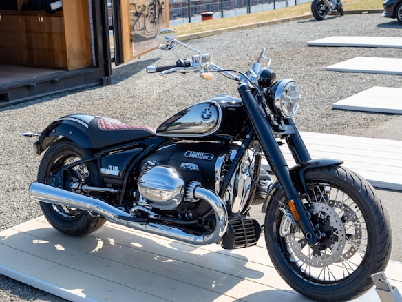 A sleek motorcycle is displayed on a light wooden platform outdoors. The motorcycle has a glossy black finish with chrome details, including the exhaust pipes and engine covers. The logo on the fuel tank is distinguishable. It is parked in an area with gravel ground and wooden platforms around, with a modern structure visible in the background.
