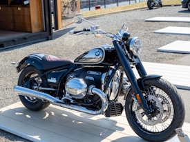 A sleek motorcycle is displayed on a light wooden platform outdoors. The motorcycle has a glossy black finish with chrome details, including the exhaust pipes and engine covers. The logo on the fuel tank is distinguishable. It is parked in an area with gravel ground and wooden platforms around, with a modern structure visible in the background.