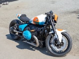 A custom motorcycle is parked on a gravel surface. The bike's design features a mix of black, white, blue, and orange colors, with sleek, robust parts and a prominent BMW logo on the gas tank and engine. It has a single seat, large exhaust pipe, and disc brakes on both wheels. The overall design is sporty and modern.