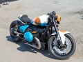A custom motorcycle is parked on a gravel surface. The bike's design features a mix of black, white, blue, and orange colors, with sleek, robust parts and a prominent BMW logo on the gas tank and engine. It has a single seat, large exhaust pipe, and disc brakes on both wheels. The overall design is sporty and modern.