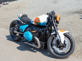 A custom motorcycle is parked on a gravel surface. The bike's design features a mix of black, white, blue, and orange colors, with sleek, robust parts and a prominent BMW logo on the gas tank and engine. It has a single seat, large exhaust pipe, and disc brakes on both wheels. The overall design is sporty and modern.