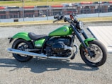 A green motorcycle is parked on a gravel surface near a racetrack. The motorcycle features a classic design with a prominent engine and chrome detailing. In the background, a chain-link fence separates the track from the viewing area with racing advertisements visible on the fence.