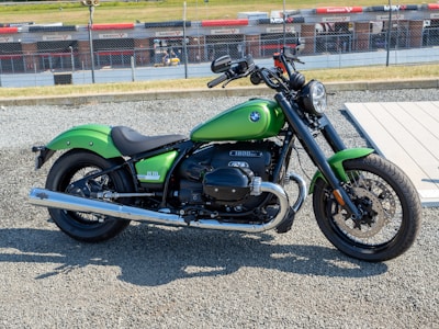 A green motorcycle is parked on a gravel surface near a racetrack. The motorcycle features a classic design with a prominent engine and chrome detailing. In the background, a chain-link fence separates the track from the viewing area with racing advertisements visible on the fence.