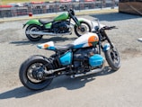 Two motorcycles are parked on a gravel surface near a race track. The motorcycle in the foreground has a distinctive custom paint job featuring white, blue, and orange accents, with visible branding on the side. The background motorbike is painted green with a classic design. Both bikes have large tires and are positioned to show off their stylish design and structure.