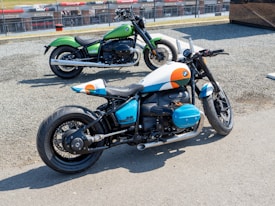 Two motorcycles are parked on a gravel surface near a race track. The motorcycle in the foreground has a distinctive custom paint job featuring white, blue, and orange accents, with visible branding on the side. The background motorbike is painted green with a classic design. Both bikes have large tires and are positioned to show off their stylish design and structure.