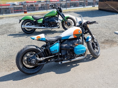Two motorcycles are parked on a gravel surface near a race track. The motorcycle in the foreground has a distinctive custom paint job featuring white, blue, and orange accents, with visible branding on the side. The background motorbike is painted green with a classic design. Both bikes have large tires and are positioned to show off their stylish design and structure.
