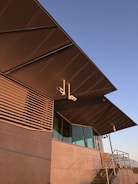 An angular modern building facade featuring large overhanging roofs, a mix of glass and metal surfaces, and external security cameras mounted near the eaves. The structure includes vertical slats and a staircase with metal railings.