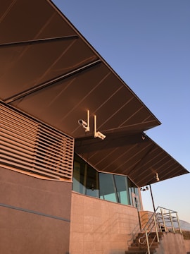 An angular modern building facade featuring large overhanging roofs, a mix of glass and metal surfaces, and external security cameras mounted near the eaves. The structure includes vertical slats and a staircase with metal railings.