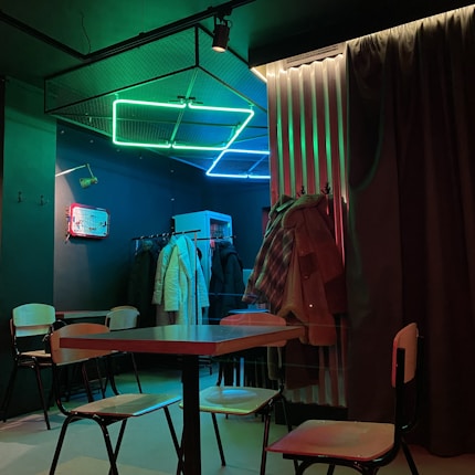 A dimly lit room with neon lights in green and blue on the ceiling. There are several coats hanging on a rack and a couple of chairs and a table positioned centrally. The walls are dark, enhancing the glow of the neon lights, and there's a curtain partially covering one side.