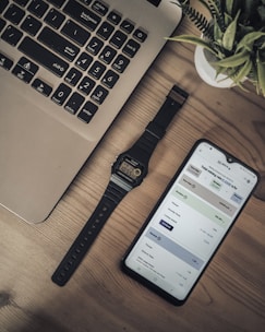 A workspace setup featuring a laptop with a visible keyboard, a digital wristwatch, and a smartphone displaying a financial or mining application on a wooden surface. A potted plant is partially visible, adding a touch of greenery to the arrangement.