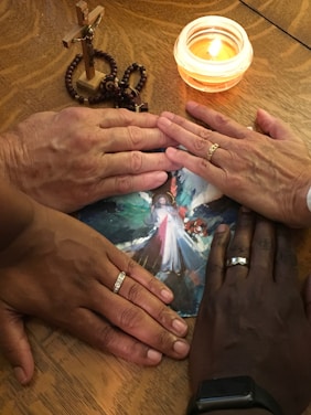 Several hands with rings are placed over a religious image on a wooden table. A wooden cross with rosary beads and a lit candle are nearby, creating a warm, intimate atmosphere.