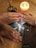 Several hands with rings are placed over a religious image on a wooden table. A wooden cross with rosary beads and a lit candle are nearby, creating a warm, intimate atmosphere.