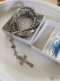 A rosary bracelet made of silver-colored beads and shells, featuring a crucifix, is displayed. The rosary is placed in an open white box with a piece of blue and white printed paper partially visible.