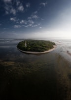 A small island with lush green vegetation surrounded by shallow, clear waters. A narrow sandy beach outlines the island, and there is a tall, white structure resembling a tower. The sky is mostly clear with a few scattered clouds, and there is an expansive view of the sea and surrounding landscape.