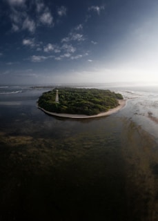A small island with lush green vegetation surrounded by shallow, clear waters. A narrow sandy beach outlines the island, and there is a tall, white structure resembling a tower. The sky is mostly clear with a few scattered clouds, and there is an expansive view of the sea and surrounding landscape.
