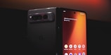 A smartphone with a foldable screen is partially unfolded, displaying its home screen with various app icons such as Keep Notes, Play Store, and Photos. The design is sleek, with a dark, reflective back and a prominent camera module. The lighting highlights the glossy surface, creating a modern and sophisticated look.