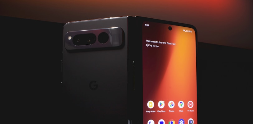 A smartphone with a foldable screen is partially unfolded, displaying its home screen with various app icons such as Keep Notes, Play Store, and Photos. The design is sleek, with a dark, reflective back and a prominent camera module. The lighting highlights the glossy surface, creating a modern and sophisticated look.