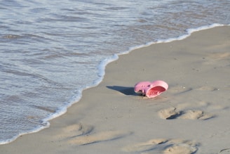 A pair of pink sandals lies abandoned on a sandy beach near the edge of gentle ocean waves. Footprints are visible in the sand nearby.