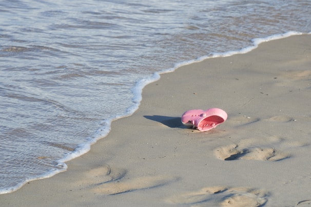 A pair of pink sandals lies abandoned on a sandy beach near the edge of gentle ocean waves. Footprints are visible in the sand nearby.