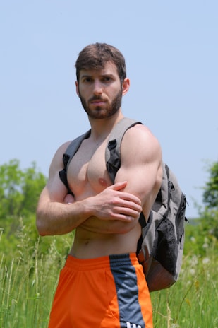 A muscular man with a beard stands shirtless in an open field, wearing bright orange shorts with black and white stripes on the sides, and a gray backpack. The man has his arms crossed over his chest and is looking directly at the camera with a serious expression. The background consists of green foliage and a clear blue sky.