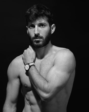 A black and white photograph of a shirtless man with a bearded face and short hair, positioned against a dark background. The man is wearing a wristwatch and is touching his neck with one hand while gazing directly at the camera.