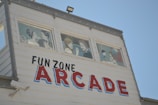 A white building with three windows displaying painted figures of people in conversation or interaction. The side of the building features the words 'FUN ZONE ARCADE' in bold, colorful letters.