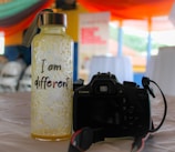 A textured bottle with a metal cap and a strap sits on a table. The bottle is labeled with the phrase 'I am different' in a playful font. Beside the bottle is a black Canon DSLR camera with a strap, positioned so the rear view is visible. The background features blurred images of chairs and tables under colorful, draped fabric, suggesting an indoor event or gathering.