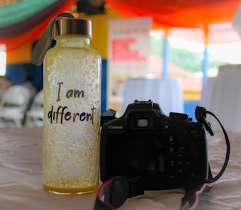 A textured bottle with a metal cap and a strap sits on a table. The bottle is labeled with the phrase 'I am different' in a playful font. Beside the bottle is a black Canon DSLR camera with a strap, positioned so the rear view is visible. The background features blurred images of chairs and tables under colorful, draped fabric, suggesting an indoor event or gathering.