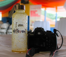 A textured bottle with a metal cap and a strap sits on a table. The bottle is labeled with the phrase 'I am different' in a playful font. Beside the bottle is a black Canon DSLR camera with a strap, positioned so the rear view is visible. The background features blurred images of chairs and tables under colorful, draped fabric, suggesting an indoor event or gathering.