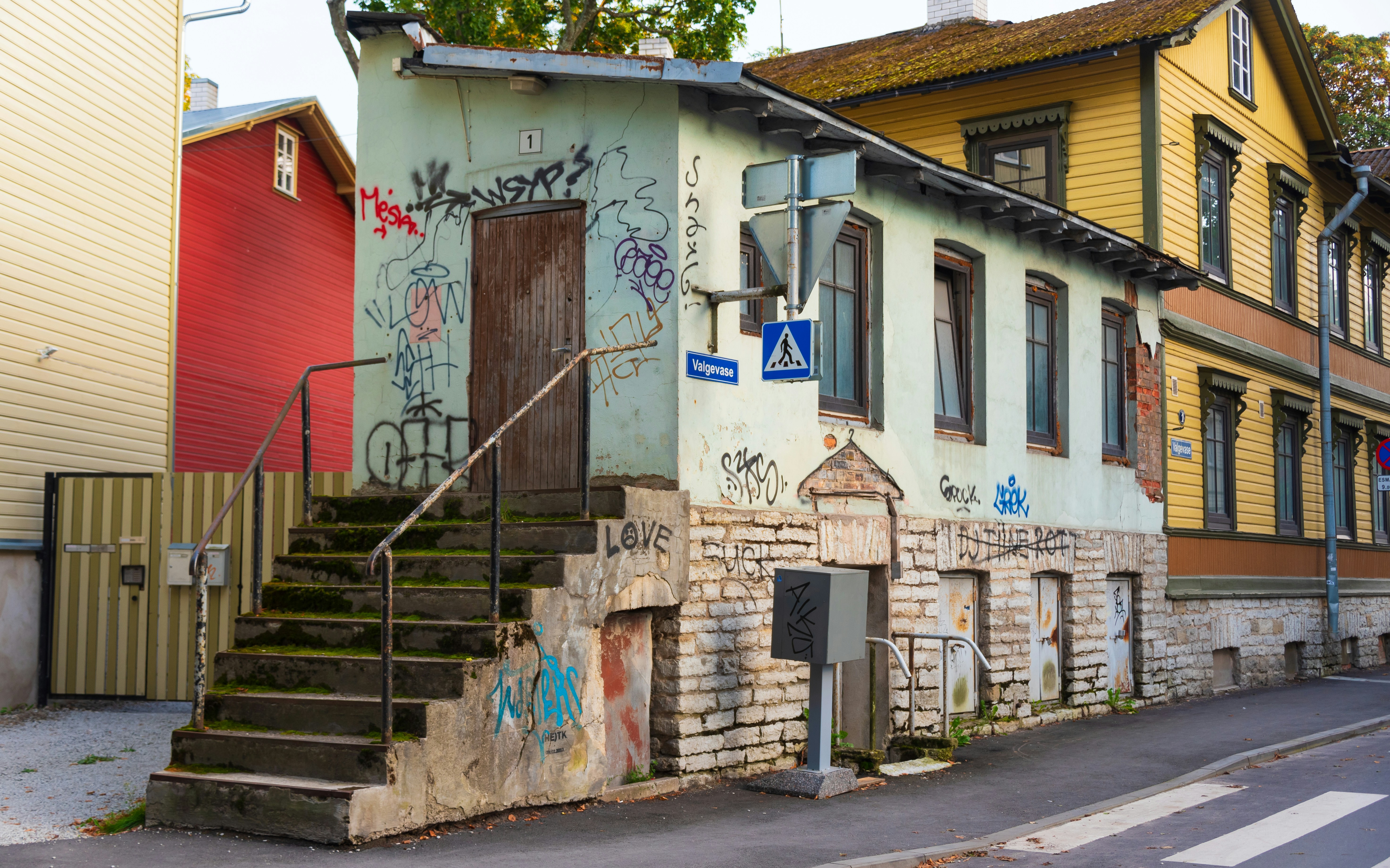 a building with graffiti on the side of it, An old building with a green exterior, covered in graffiti, stands at the corner. The building has a wooden door at the top of a mossy staircase and several windows. Surrounding buildings are painted in different colors, adding to the vibrant atmosphere of the area. A pedestrian crossing sign is visible, indicating a crosswalk nearby.