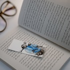 An open book with text written in Persian script, accompanied by a cartoonish bookmark depicting a person wearing a blue jacket and helmet sitting on a scooter. In the background, a pair of glasses rests on the white surface.