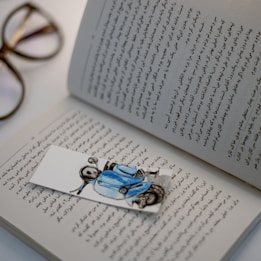 An open book with text written in Persian script, accompanied by a cartoonish bookmark depicting a person wearing a blue jacket and helmet sitting on a scooter. In the background, a pair of glasses rests on the white surface.
