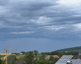 Overcast sky with heavy, layered clouds looming above a rural landscape. The scene includes a field with rows of trees and a construction crane in the distance. In the foreground, solar panels are visible on a rooftop, indicating a mix of natural and developed environments.