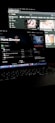 A dimly lit room featuring a laptop in the foreground, open to a music streaming service displaying Hans Zimmer's profile. In the background, a larger screen shows a YouTube interface with various video thumbnails. Both screens have a dark mode theme.