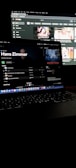A dimly lit room featuring a laptop in the foreground, open to a music streaming service displaying Hans Zimmer's profile. In the background, a larger screen shows a YouTube interface with various video thumbnails. Both screens have a dark mode theme.