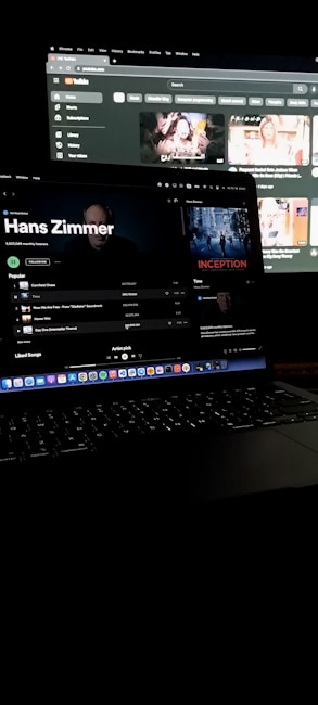 A dimly lit room featuring a laptop in the foreground, open to a music streaming service displaying Hans Zimmer's profile. In the background, a larger screen shows a YouTube interface with various video thumbnails. Both screens have a dark mode theme.