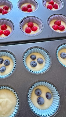 A tray of uncooked muffins in cupcake liners, each filled with batter and topped with either raspberries or blueberries. The liners are a mix of light blue and purple, arranged neatly in a muffin tin.