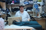 A man is sitting in a room surrounded by shelves filled with neatly folded fabrics. In front of him is a sewing machine with some material partially sewn under its foot. To the right of the man, there is an electric fan on a table. The setting appears to be a small tailor shop or workshop.