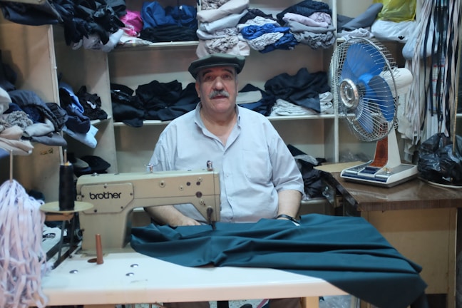 A man is sitting in a room surrounded by shelves filled with neatly folded fabrics. In front of him is a sewing machine with some material partially sewn under its foot. To the right of the man, there is an electric fan on a table. The setting appears to be a small tailor shop or workshop.