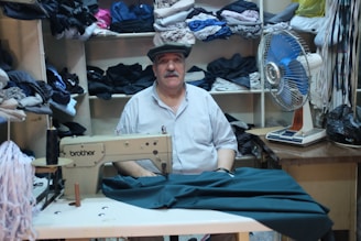 A man is sitting in a room surrounded by shelves filled with neatly folded fabrics. In front of him is a sewing machine with some material partially sewn under its foot. To the right of the man, there is an electric fan on a table. The setting appears to be a small tailor shop or workshop.